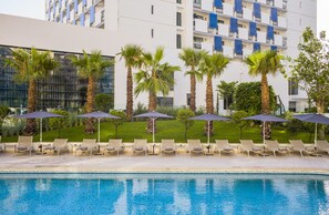 A swimming pool with lounge chairs and umbrellas, surrounded by palm trees and a modern building with balconies.