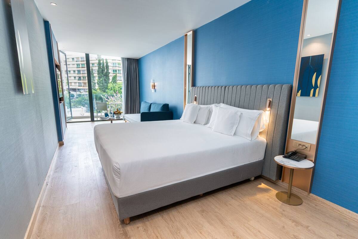 A modern hotel room with a large bed, a bedside table with a lamp, a small round table, and a large mirror.