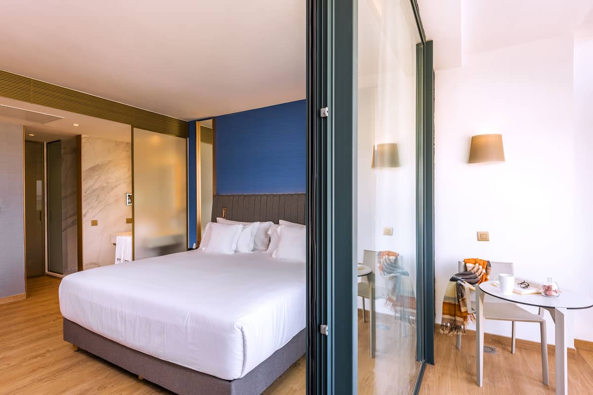 A modern hotel room with a large bed, a glass door leading to a balcony, a small table with a chair, and a wall-mounted lamp.