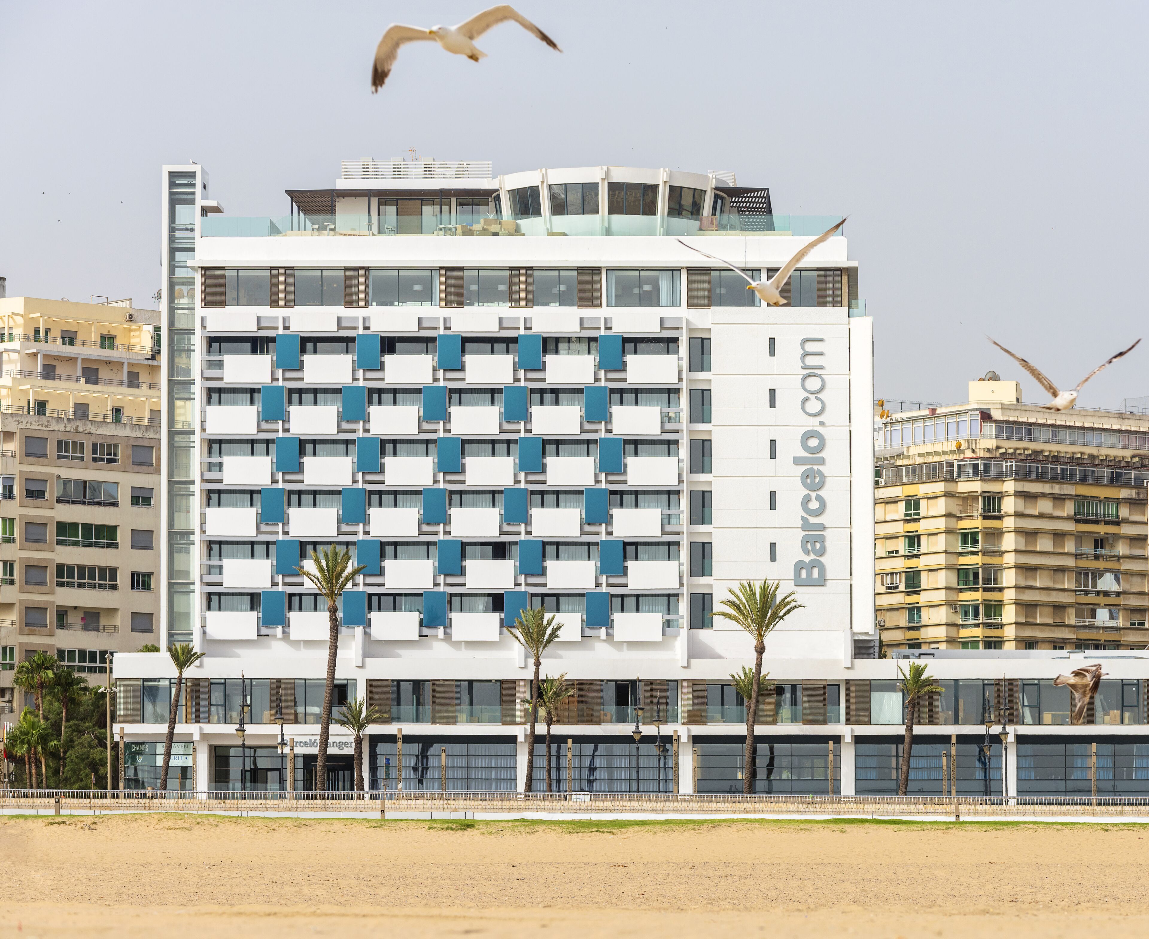 Un hotel moderno con una serie de balcones, un cartel llamativo del sitio web y palmeras en primer plano.