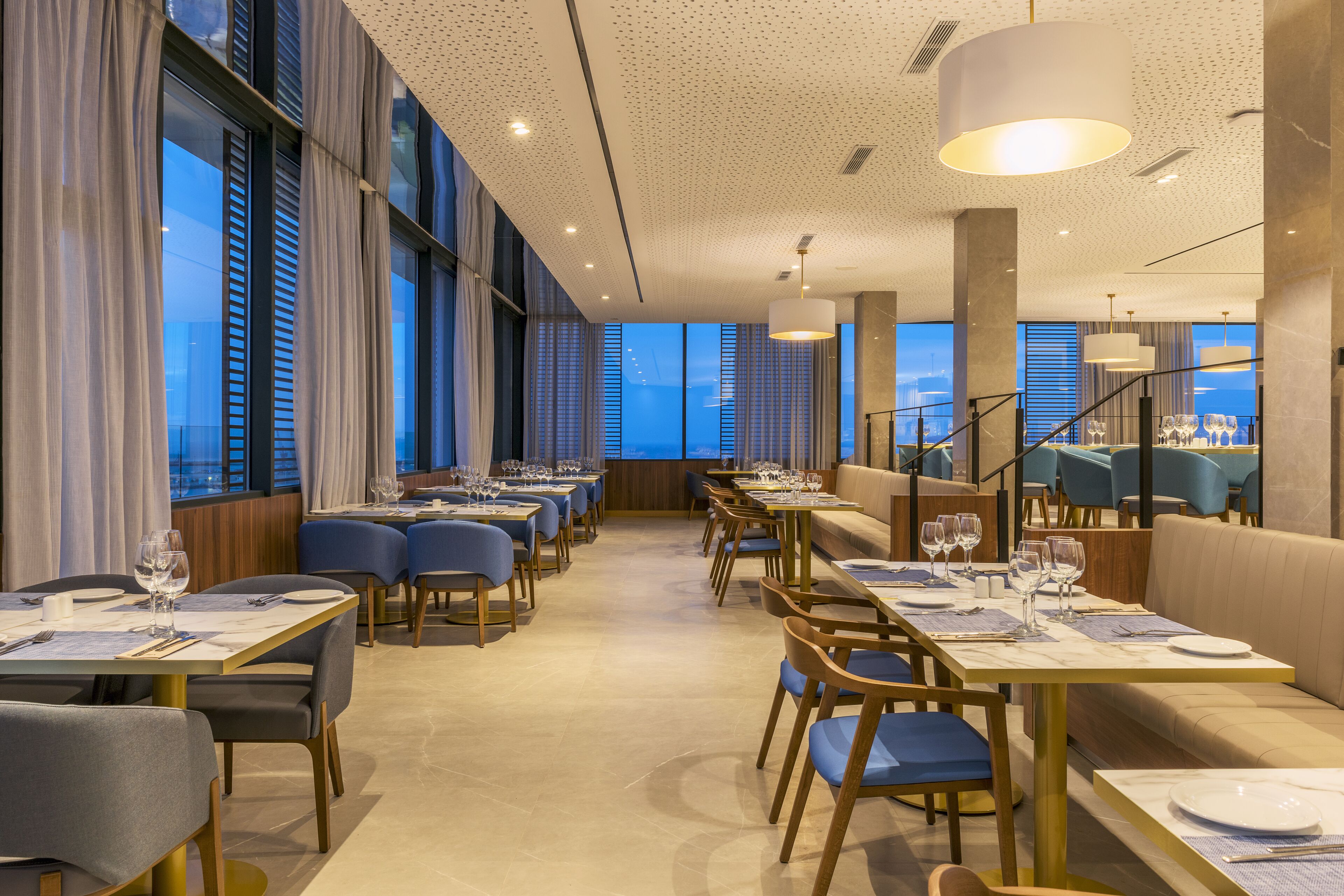 A modern restaurant with long tables, blue chairs, and a view of the cityscape.