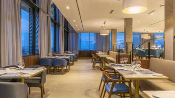 A modern restaurant with long tables, blue chairs, and a view of the cityscape.