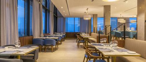 A modern restaurant with long tables, blue chairs, and a view of the cityscape.
