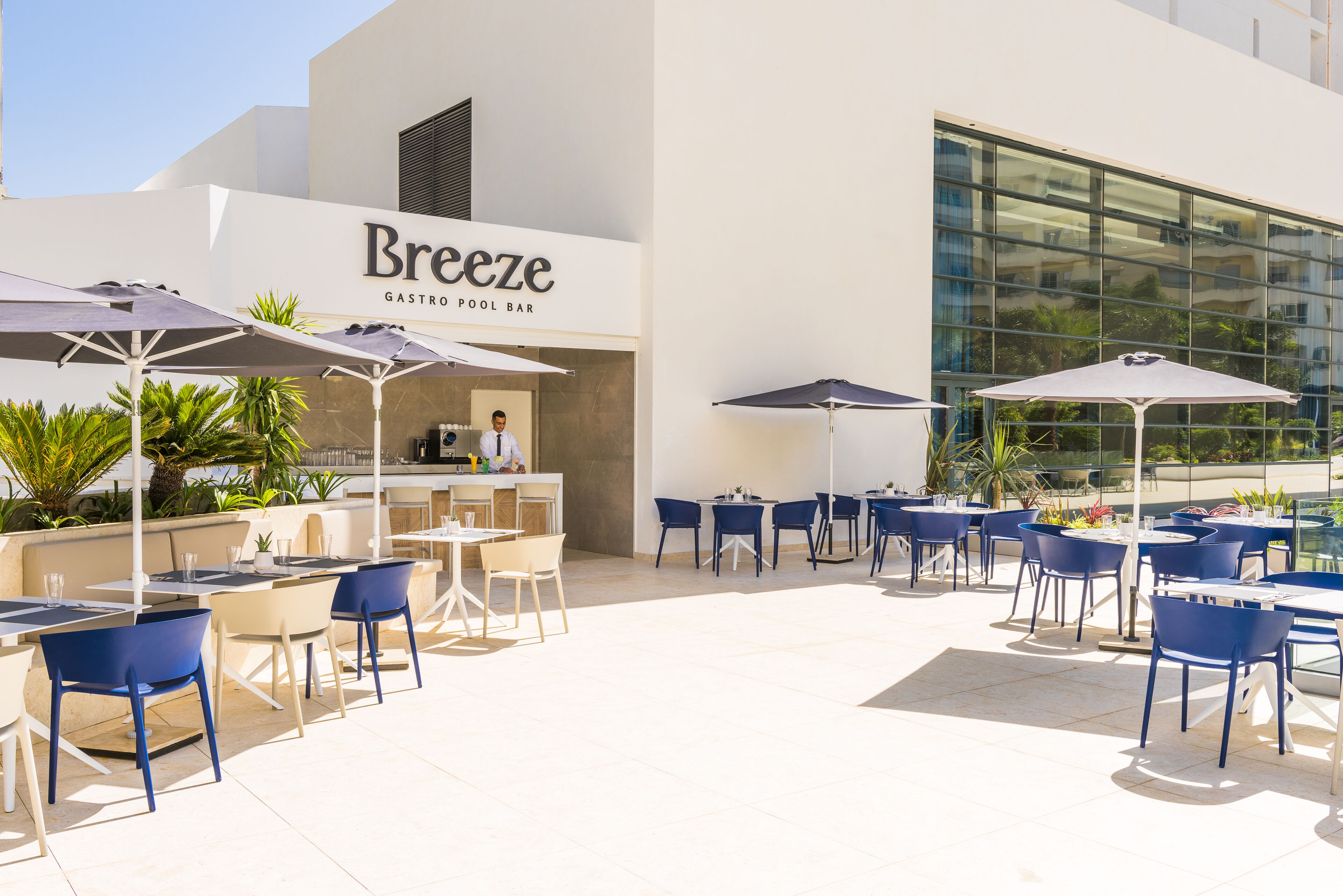 Un área de comedor al aire libre con mesas, sillas, sombrillas y un bar junto a la piscina llamado 'Breeze'.