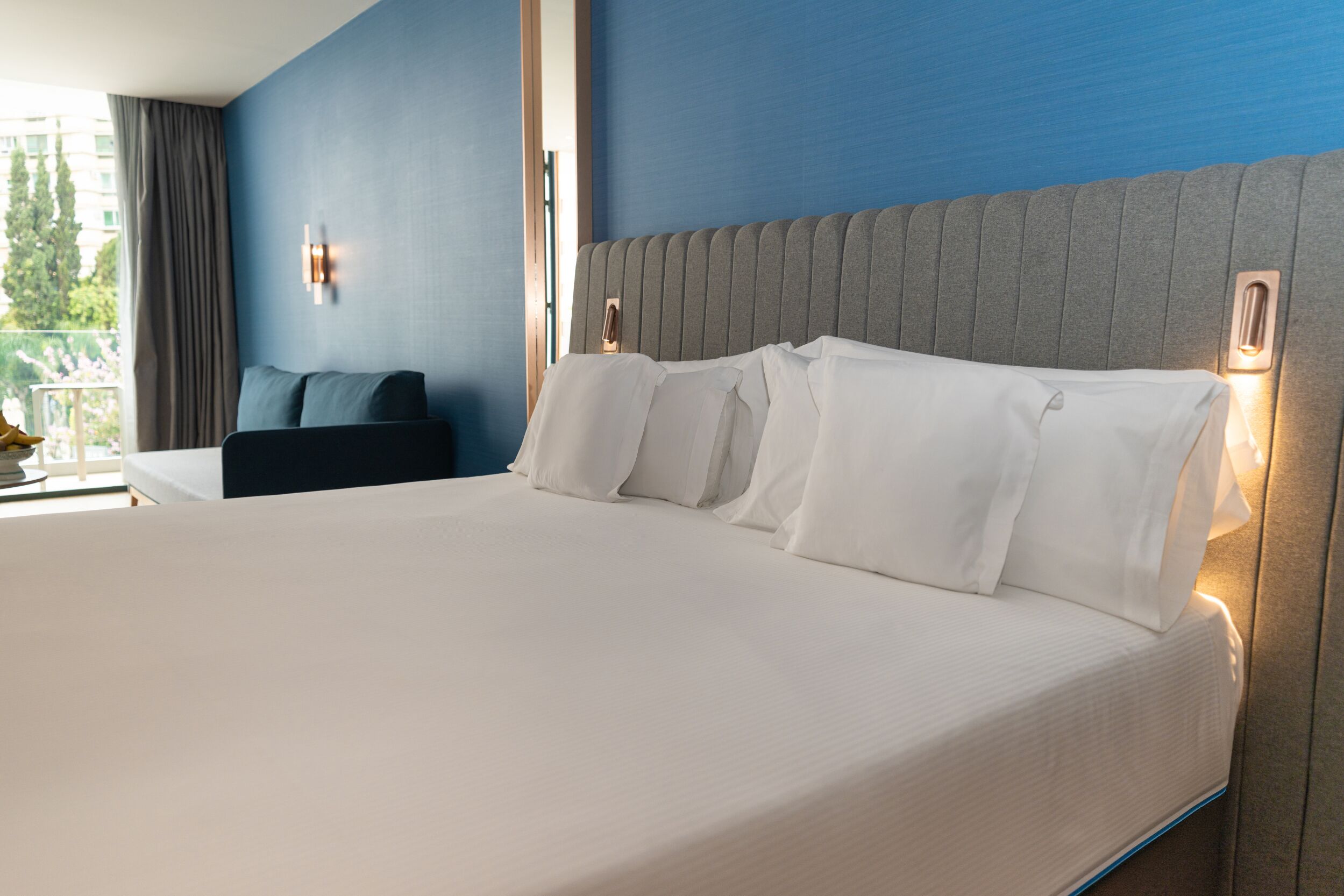 A hotel room with a large bed, a headboard with built-in lighting, a dark-colored armchair, and a window with curtains.