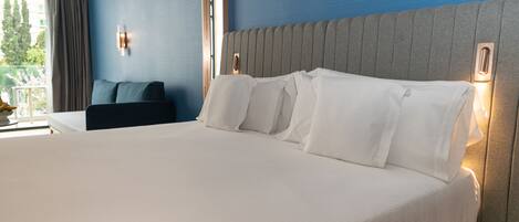 A hotel room with a large bed, a headboard with built-in lighting, a dark-colored armchair, and a window with curtains.