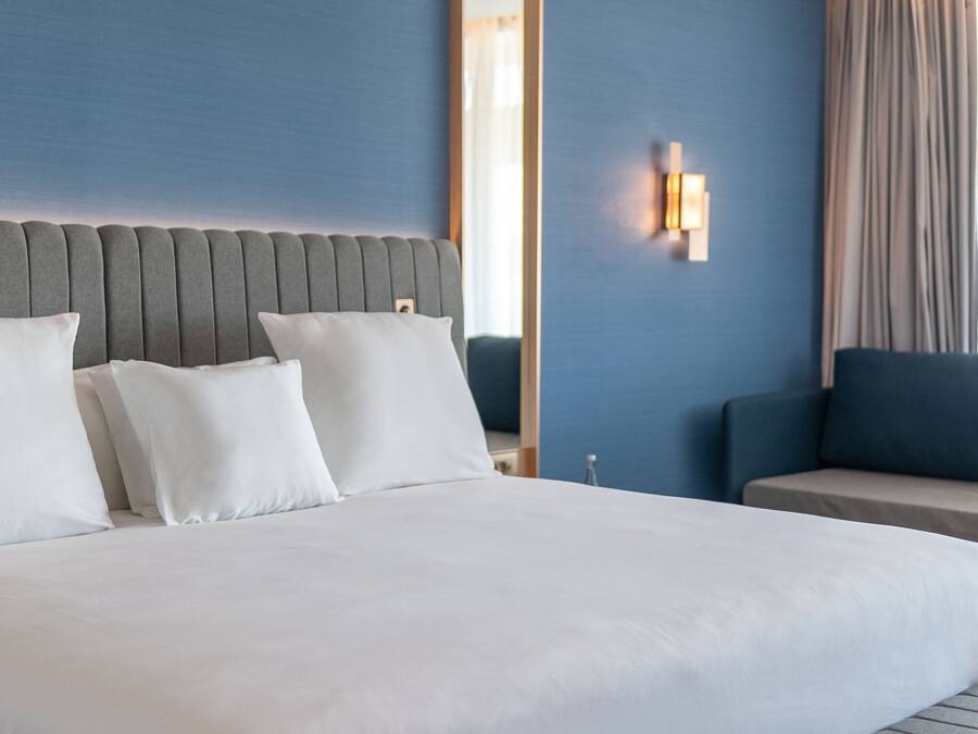 Una cama bien hecha con sábanas y almohadas blancas, un cabecero tapizado en gris, una lámpara de pared y vistas a una ventana con cortinas.