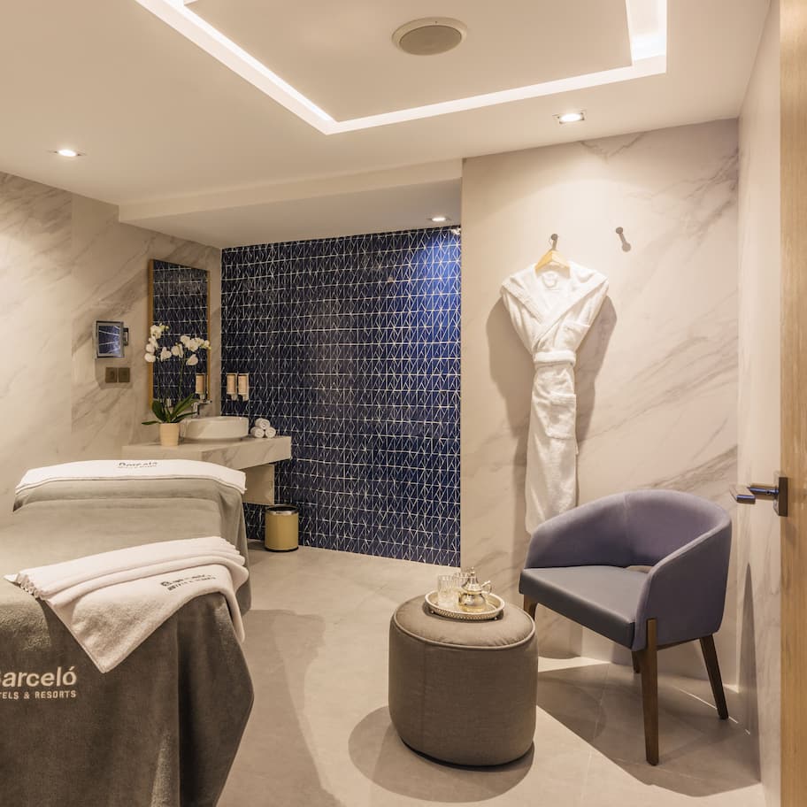 A modern spa room with two massage tables, a small seating area, and a blue patterned wall.