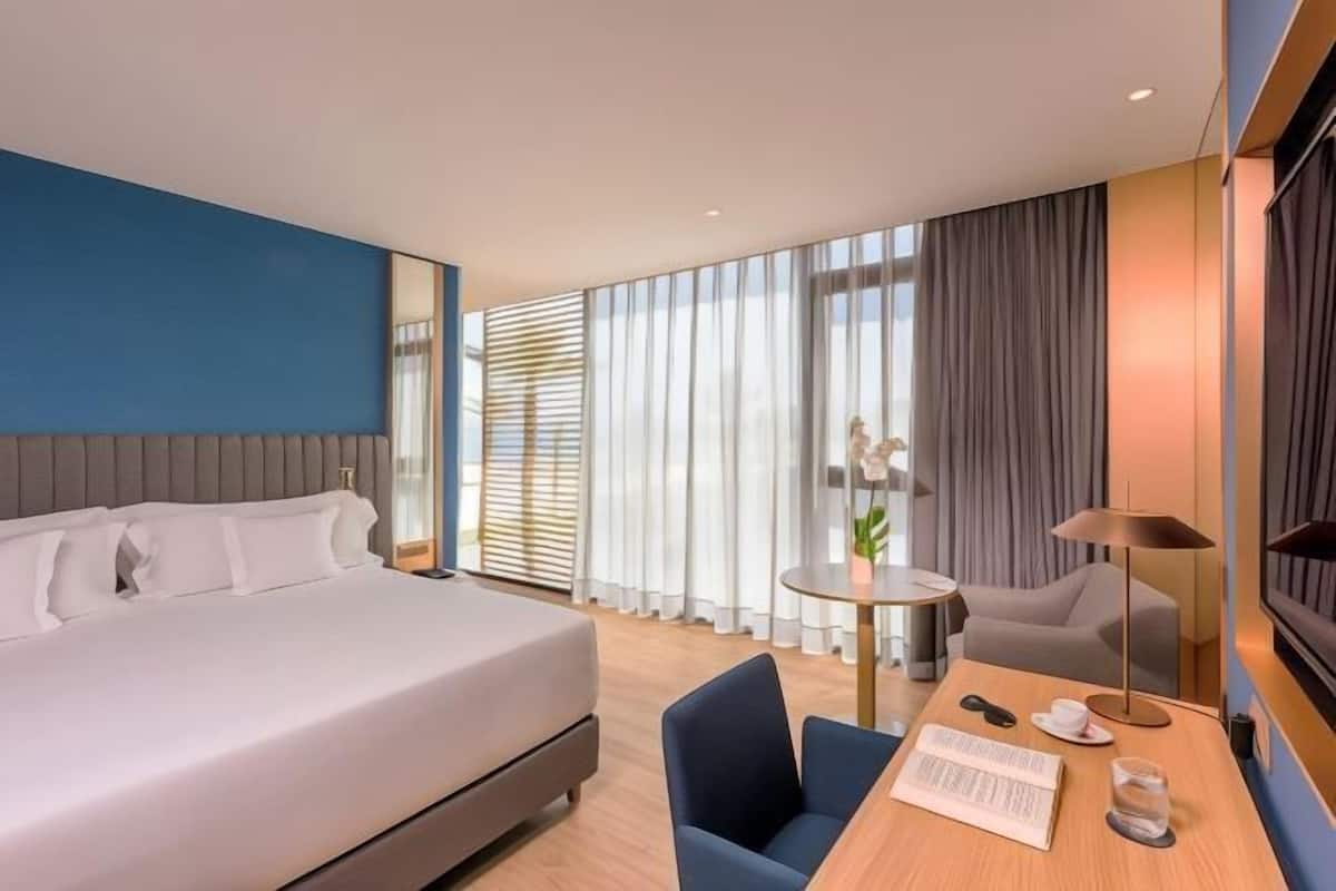 A modern hotel room with a large bed, a desk with a chair, a small table, and a lamp.