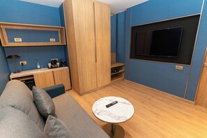 A compact room with a sofa, a small round table with a remote control, a TV mounted on the wall, and a wooden cabinet.