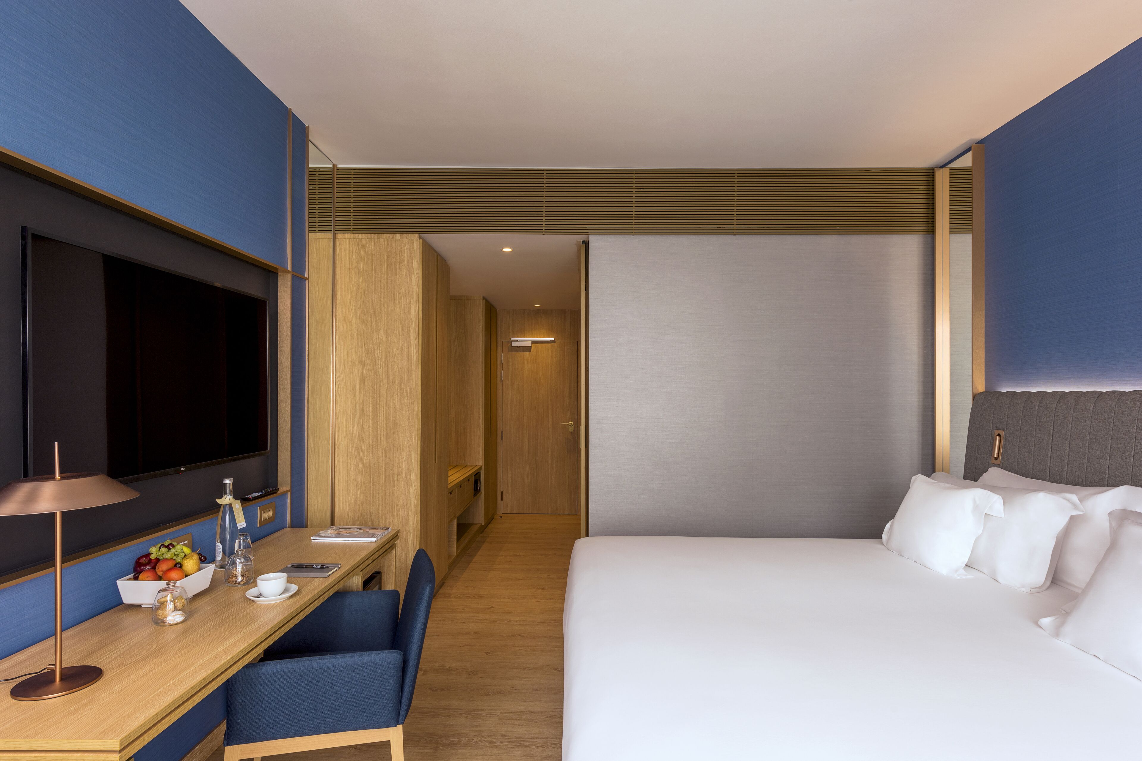 A modern hotel room with a large bed, a desk with a flat-screen TV, a chair, and a small table with a fruit bowl.