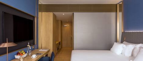 A modern hotel room with a large bed, a desk with a flat-screen TV, a chair, and a small table with a fruit bowl.