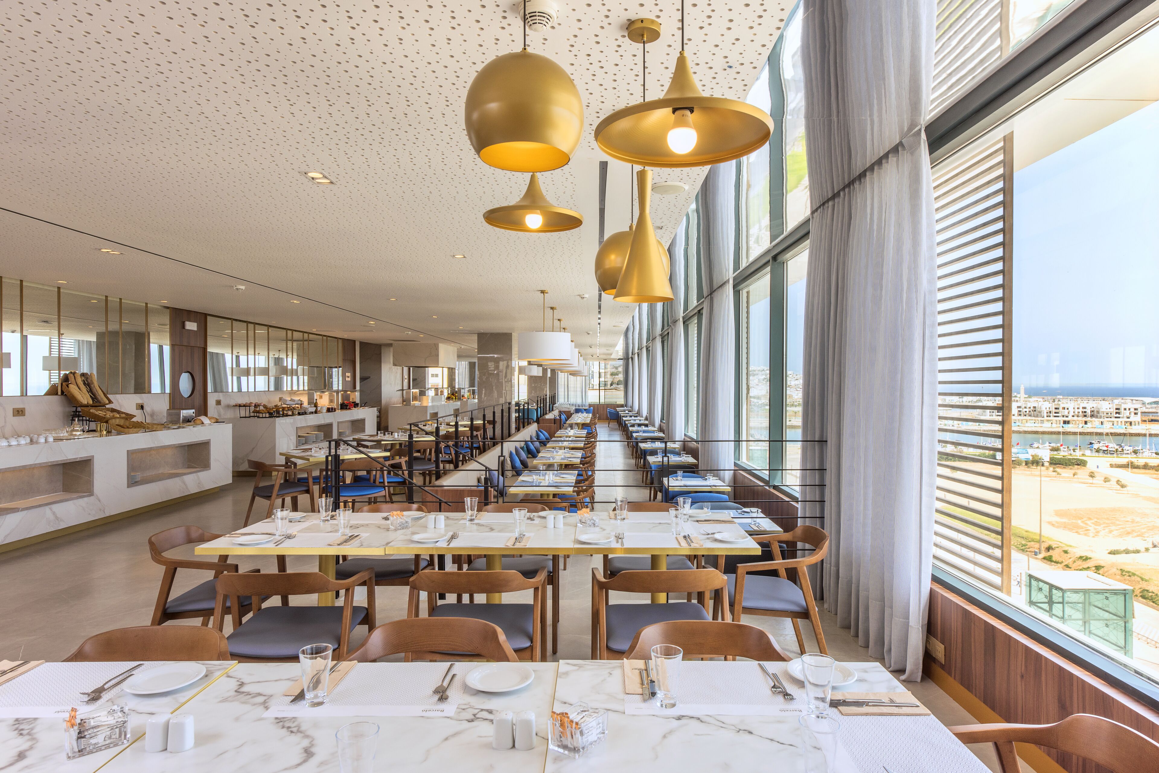 A spacious dining area with long tables and chairs, large windows offering a view of the beach, and modern pendant lighting.