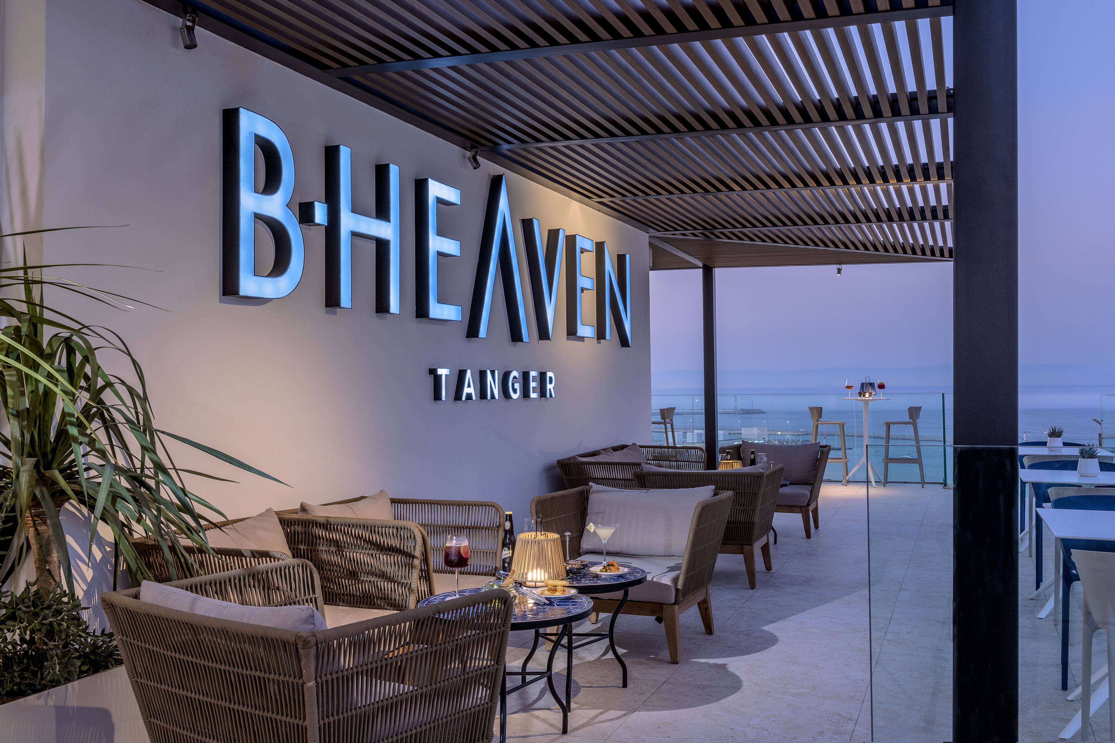 A rooftop bar with wicker furniture, a table with a drink, and a view of the sea.