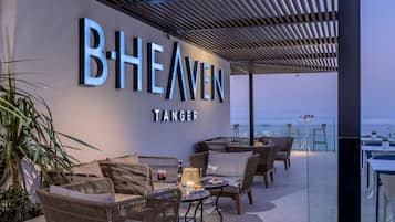 A rooftop bar with wicker furniture, a table with a drink, and a view of the sea.