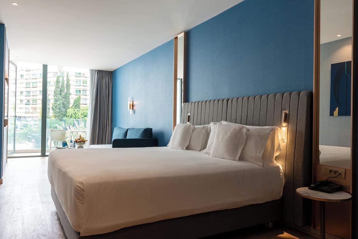 A modern hotel room with a large bed, a bedside table with a phone, and a view of a building and trees through the window.