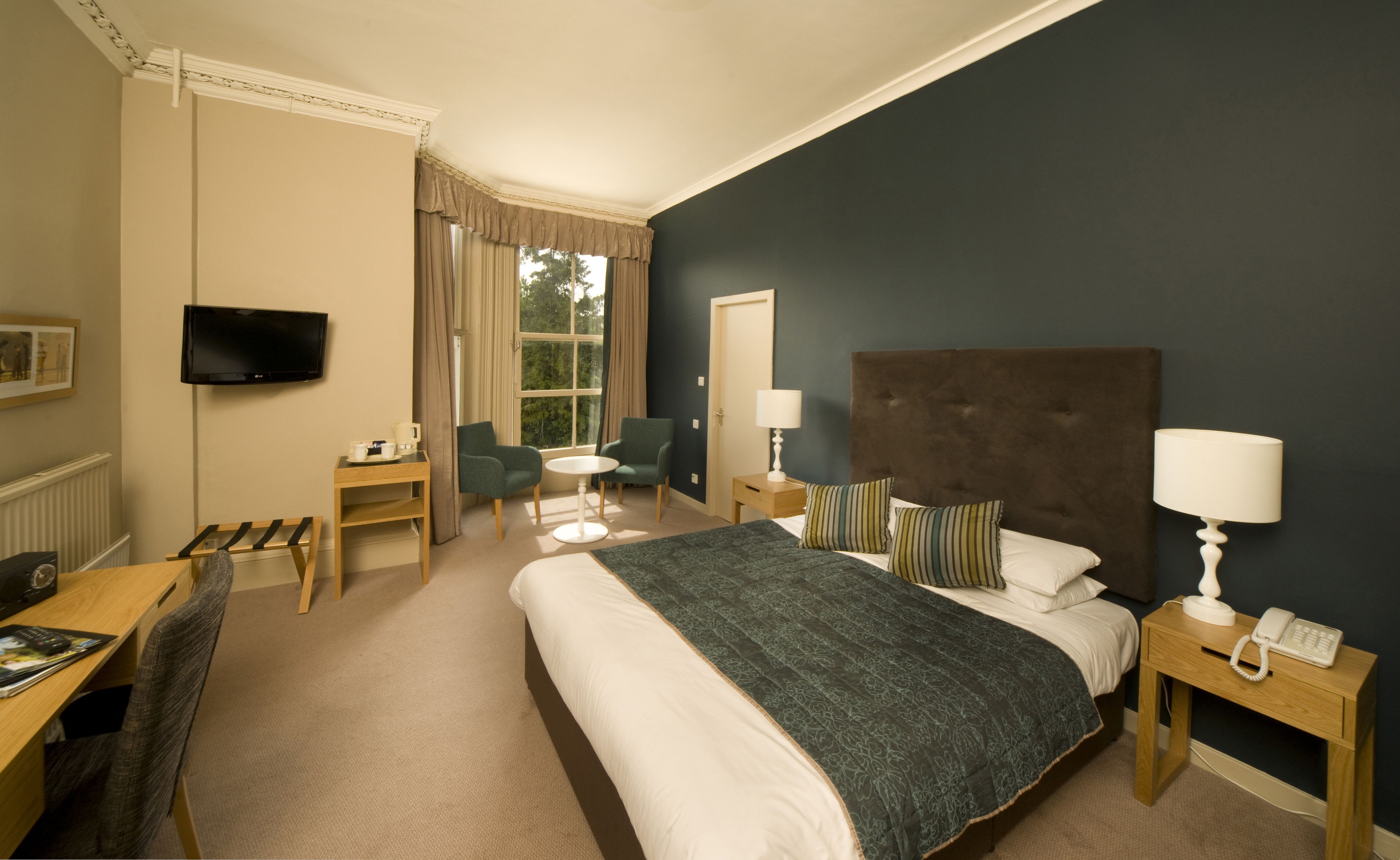 Habitación de hotel con una cama grande, dos mesitas de noche, un escritorio con silla, un televisor montado en la pared y una ventana con cortinas.
