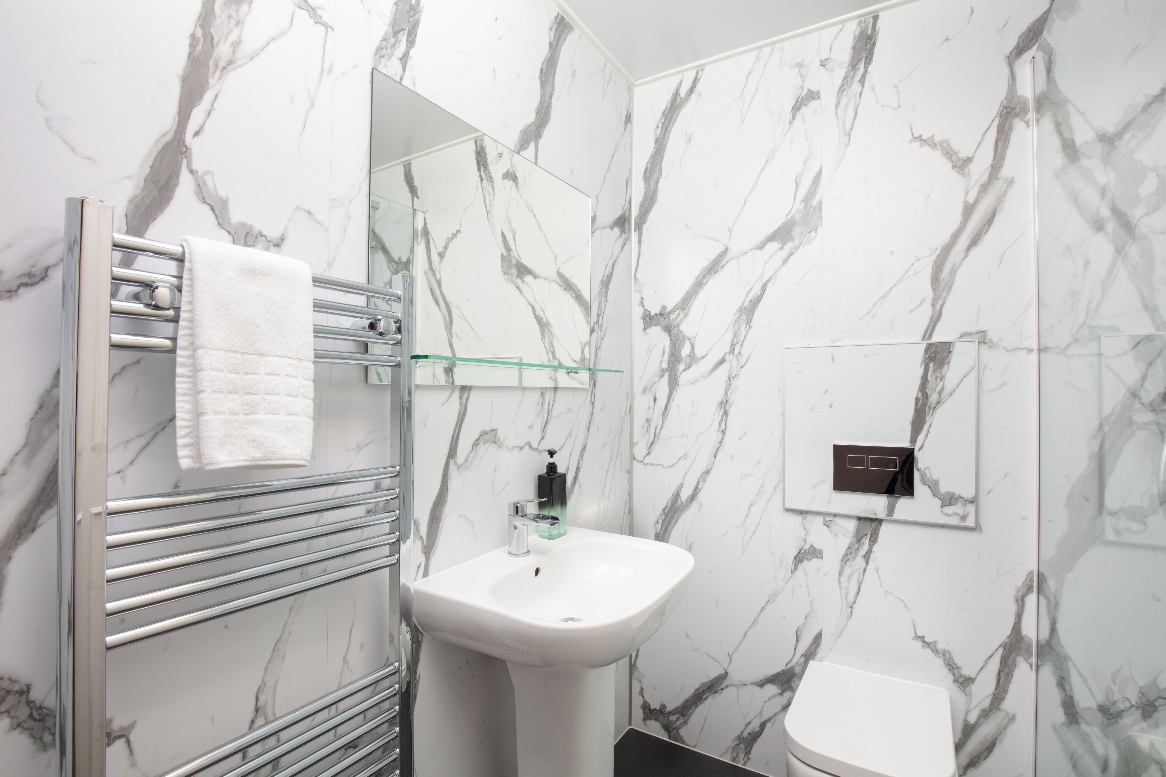 Een badkamer met een marmeren wand, een glazen douchecabine, een wit wastafeltje en een handdoekhouder.