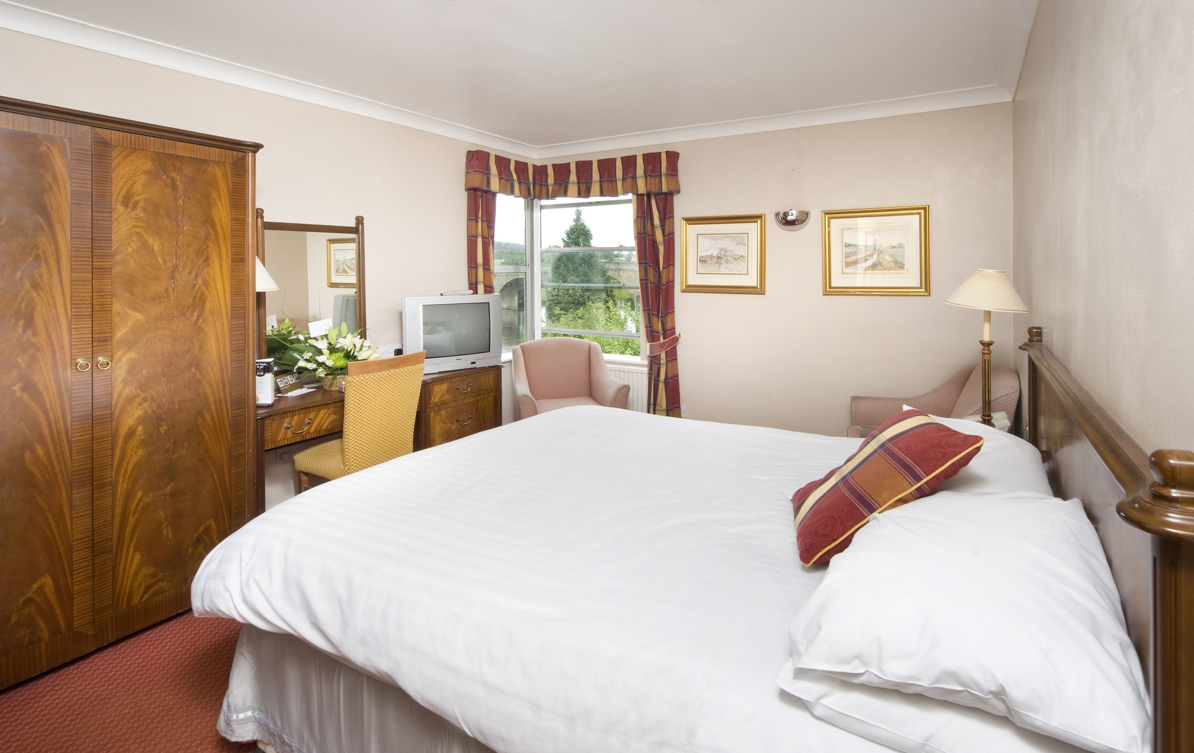 A hotel room with a large bed, a wooden wardrobe, a desk with a television, a chair, and a window with curtains.