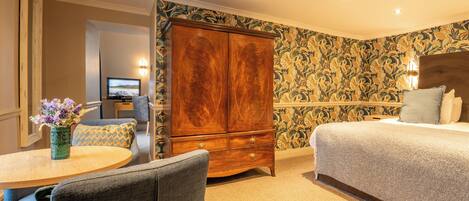 A hotel room with a bed, a wooden wardrobe, a round table with a vase, a television, and a patterned wallpaper.