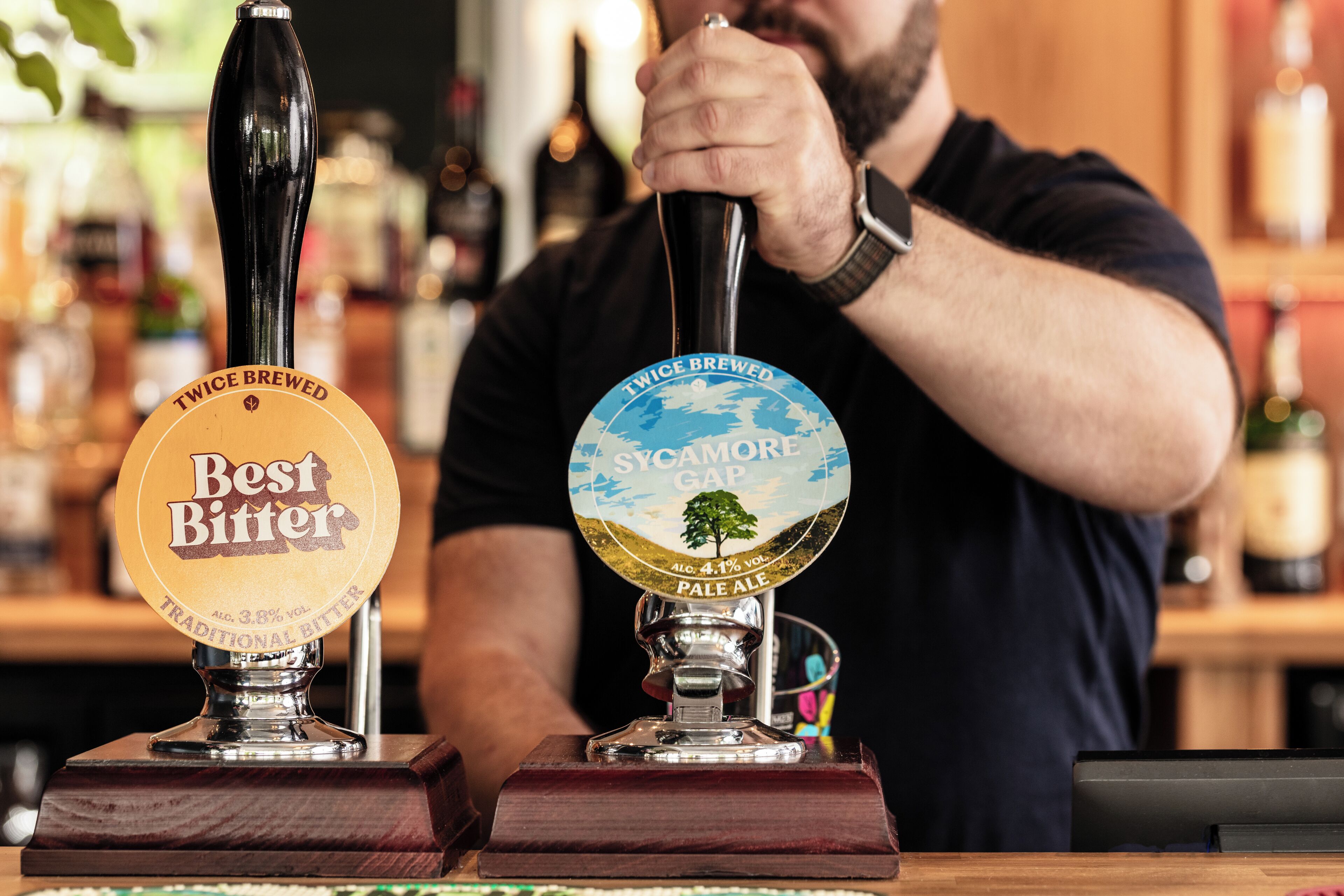 A bartender pouring beer from two taps labeled "Best Bitter" and "Sycamore Gap Pale Ale."