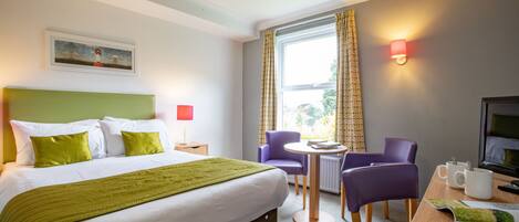 Habitación de hotel con cama, escritorio, dos sillas, mesa, lámpara, televisor y ventana con cortinas.