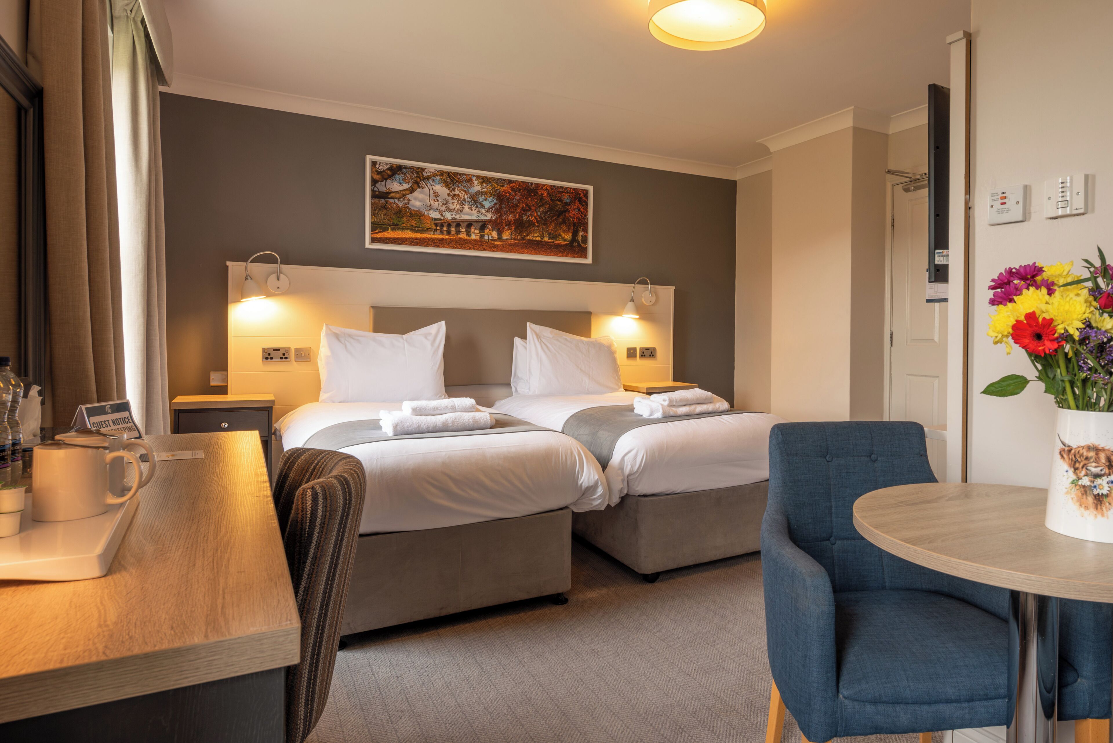 A hotel room with two beds, a desk, a chair, a table with a vase of flowers, and a painting on the wall.