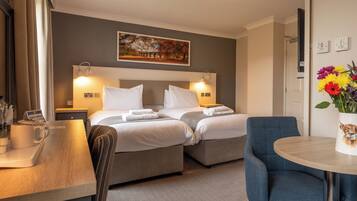 A hotel room with two beds, a desk, a chair, a table with a vase of flowers, and a painting on the wall.