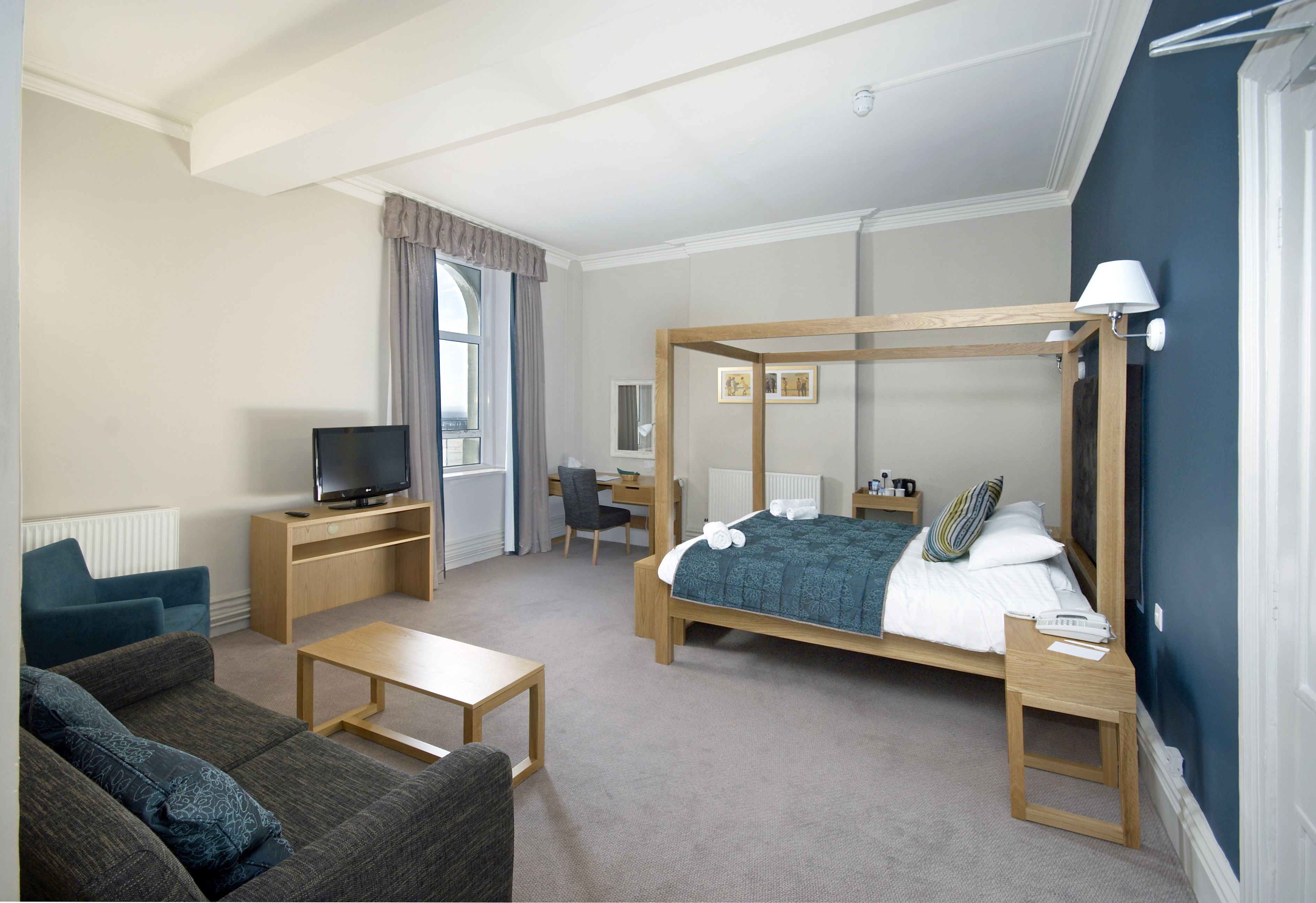 A modern hotel room with a four-poster bed, a desk with a computer, a television, a sofa, and a window with curtains.