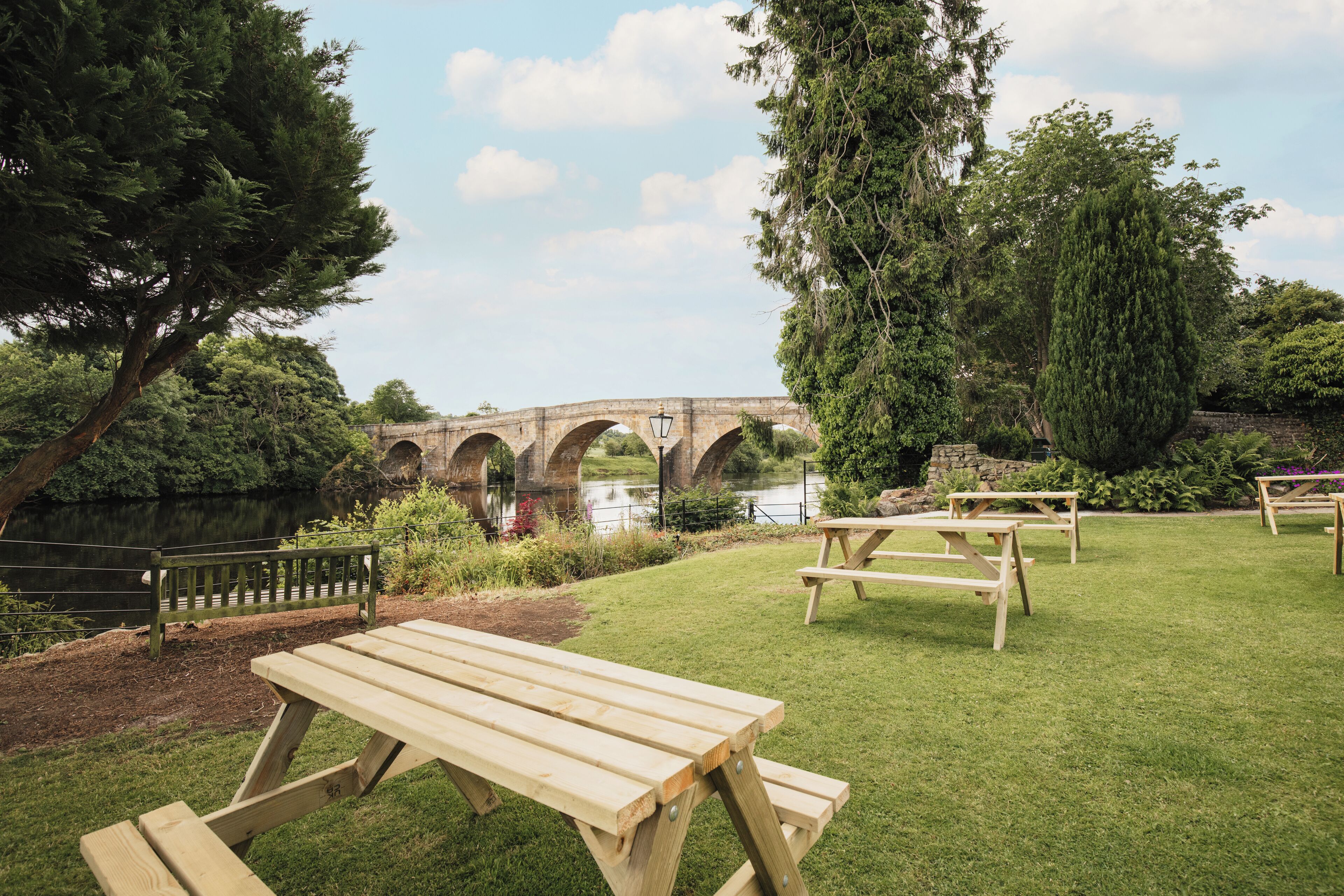 Ein Picknickplatz mit Holztischen und -bänken, im Hintergrund eine Steinbrücke und ein Gewässer.