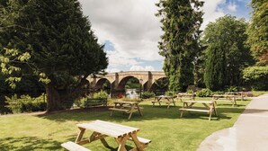 Un área de picnic con mesas y bancos de madera, un puente de piedra y abundante vegetación.