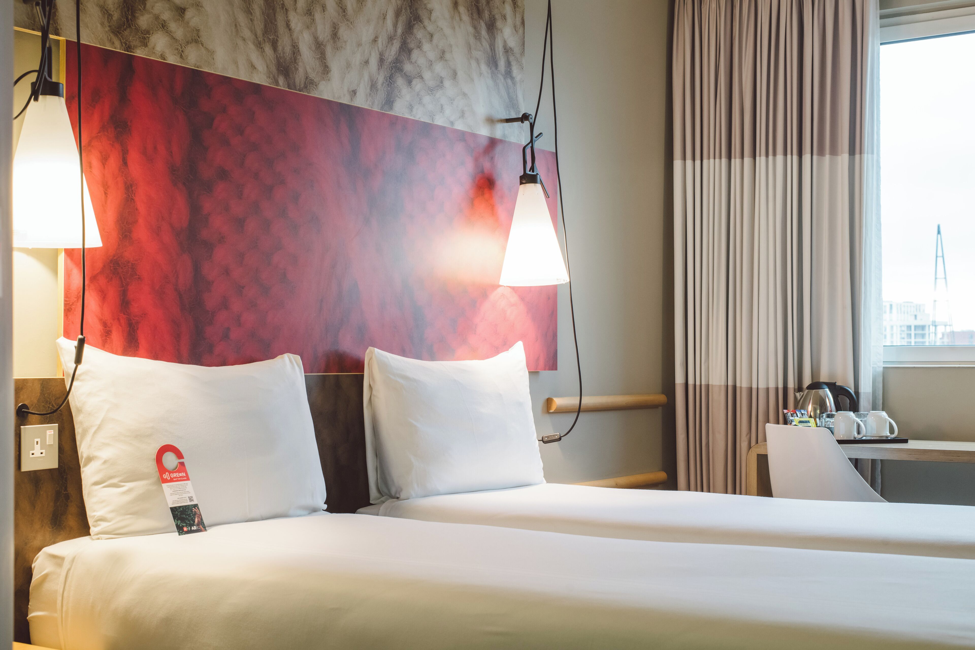 A hotel room with a bed, two bedside lamps, a nightstand, a desk, and a window with curtains.