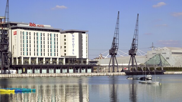 Un hotel moderno con un llamativo letrero de 'ibis HOTEL', un letrero de 'NOVOTEL' y una estructura de grúa al fondo.