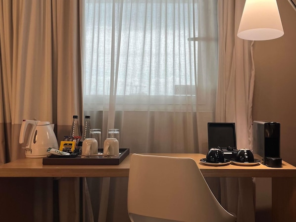 Une chambre d’hôtel équipée d’un bureau, d’une chaise, d’une machine à café, d’une bouilloire, d’un plateau avec des condiments et d’une fenêtre avec des rideaux.