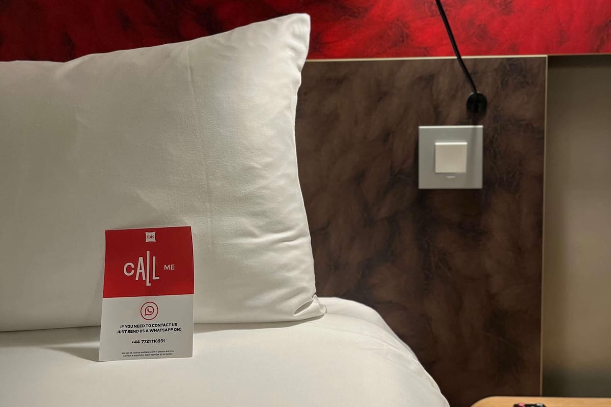 Ett hotellrum med en vit kudde, en röd och vit skylt med texten ”RING MIG”, ett eluttag och en ljusströmbrytare på väggen.