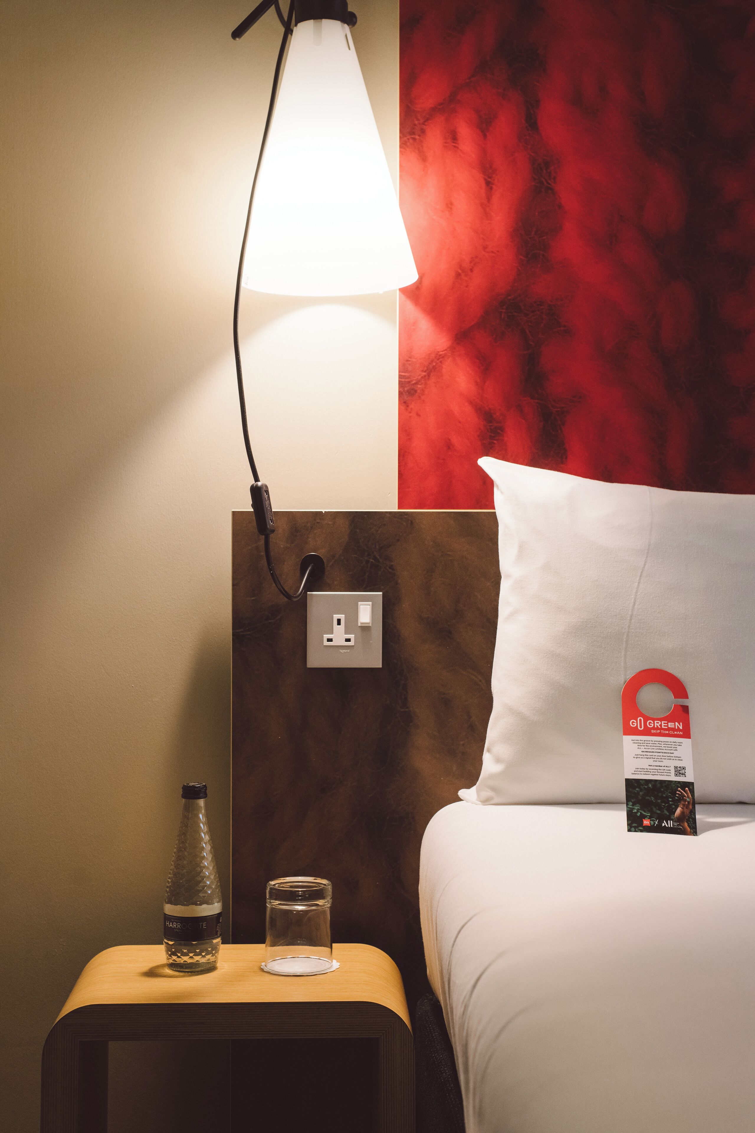 Hotellihuone, jossa on sänky, yöpöytä lampun kanssa, pullo ja lasi.