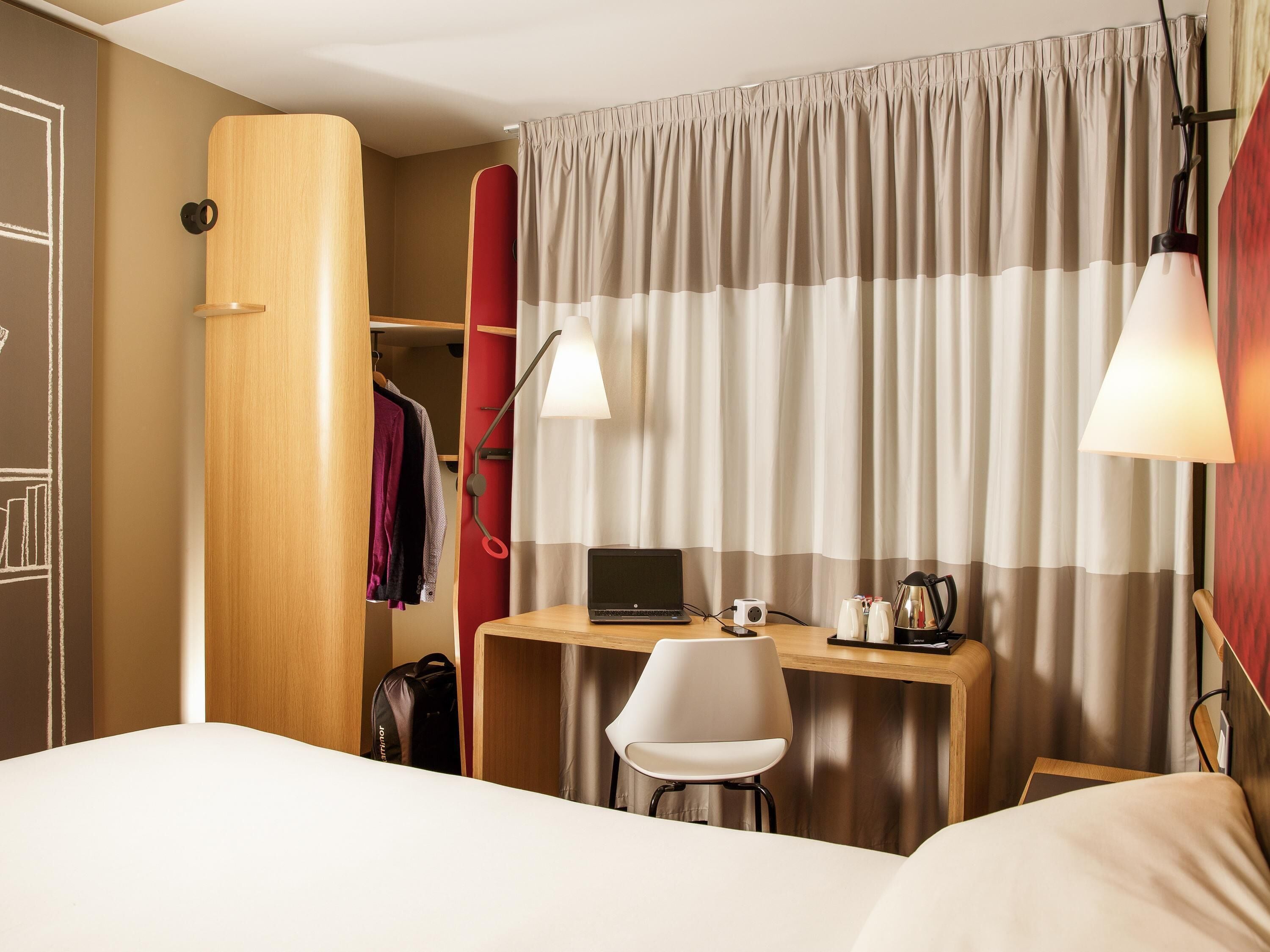 A hotel room with a bed, a desk with a laptop, a chair, a wardrobe, and a curtain.