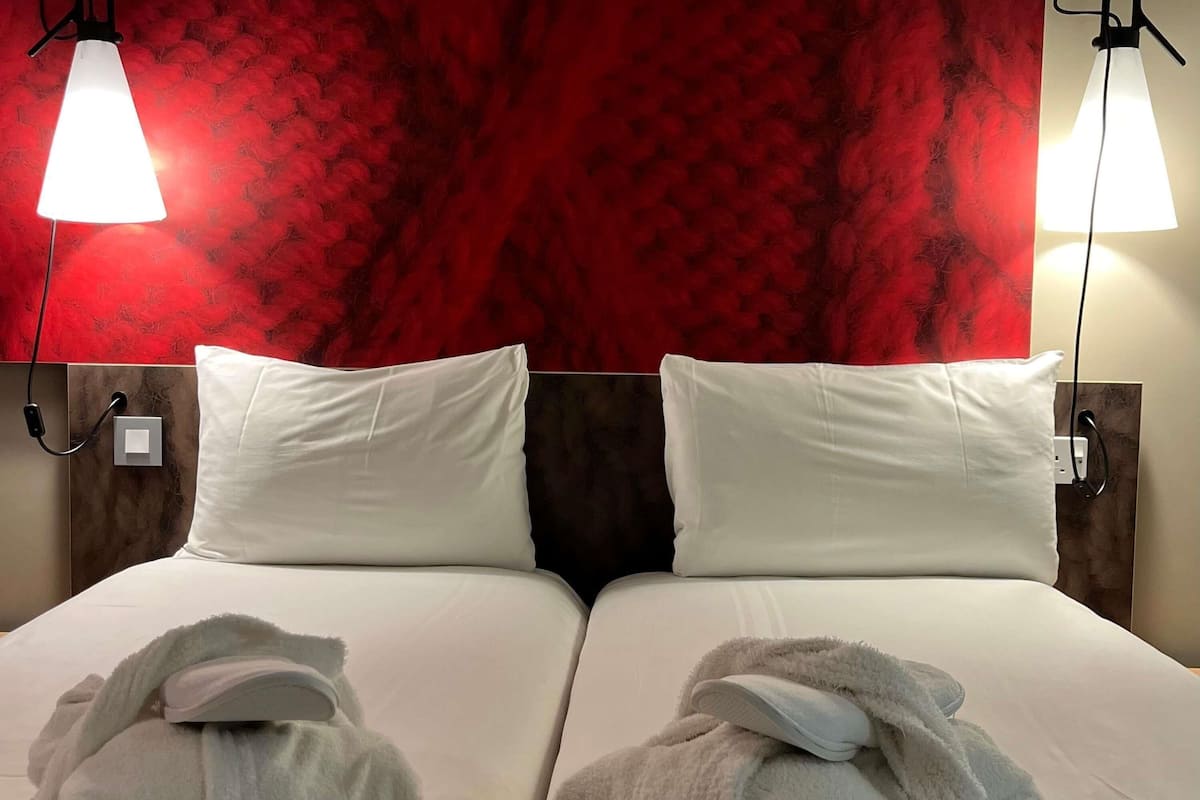 Ett hotellrum med två sängar, vita sängkläder, en röd vägg med struktur och två sänglampor.