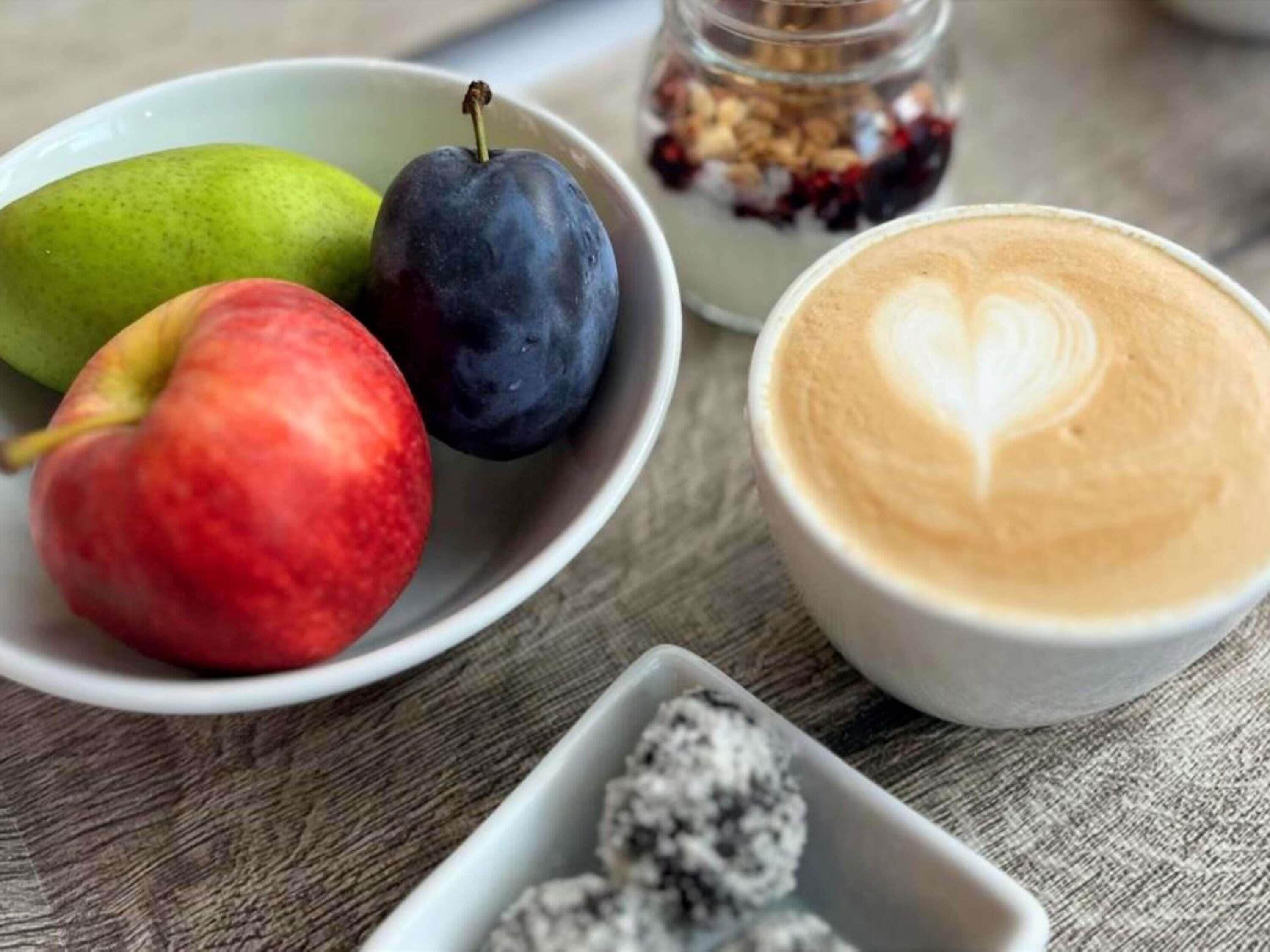 Una mesa con un bol de fruta, un tarro de granola, una taza de café con arte latte y un recipiente pequeño de bayas.