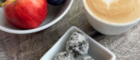 Una mesa con un bol de fruta, un tarro de granola, una taza de café con arte latte y un recipiente pequeño de bayas.