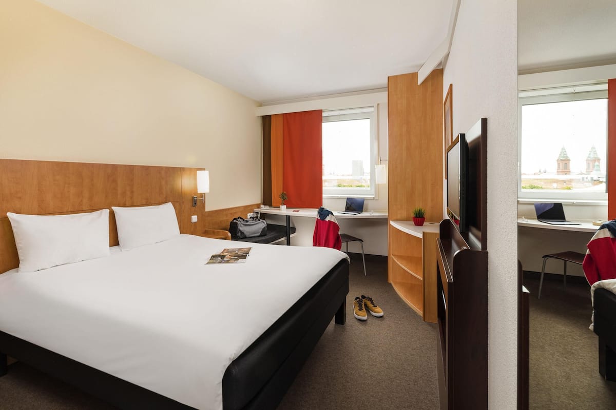 A hotel room with a large bed, a desk with a laptop, a TV, and a view of a building through the window.