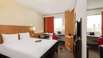 A hotel room with a large bed, a desk with a laptop, a TV, and a view of buildings through the window.