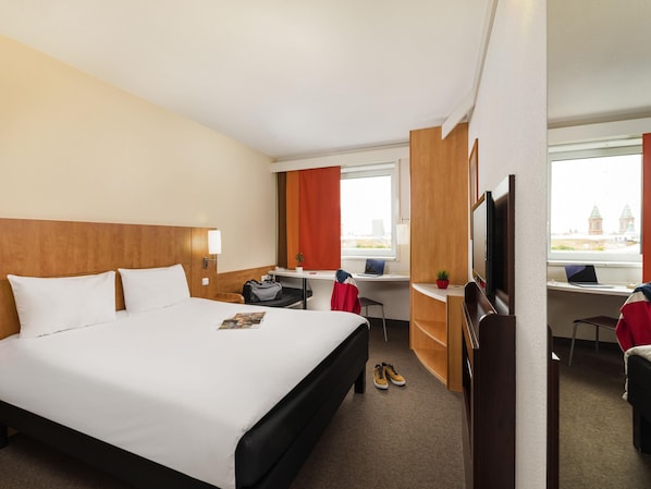 A hotel room with a large bed, a desk with a laptop, a TV, and a view of buildings through the window. - ibis Praha Mala Strana (Prague)