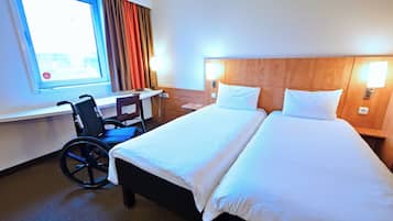 Habitació d'hotel amb cadira de rodes, escriptori, llit amb roba blanca i finestra amb cortines.