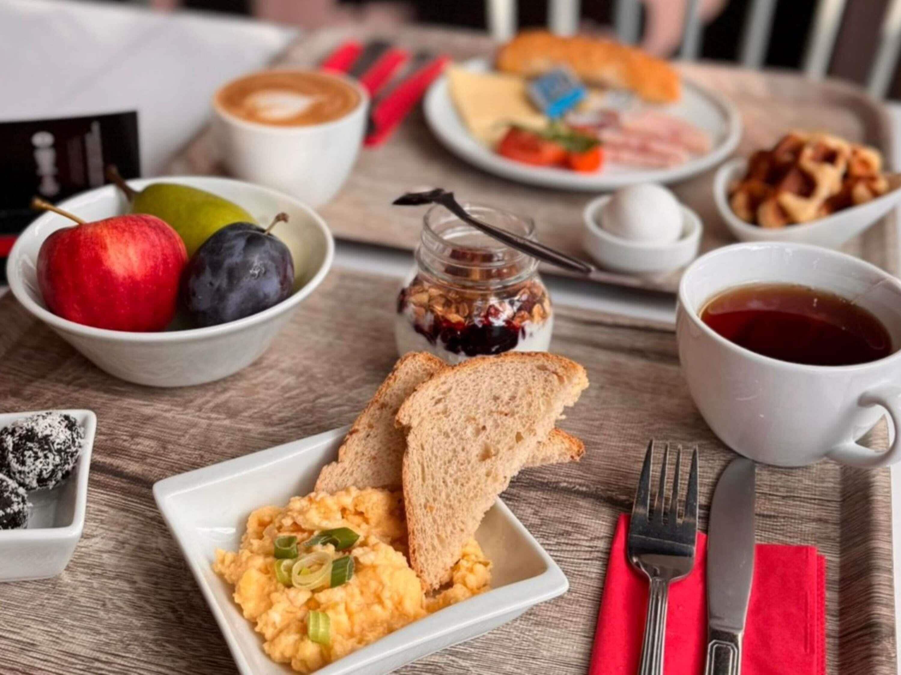 A breakfast tray with toast, scrambled eggs, a bowl of fruit, a cup of coffee, and a plate with ham and cheese.