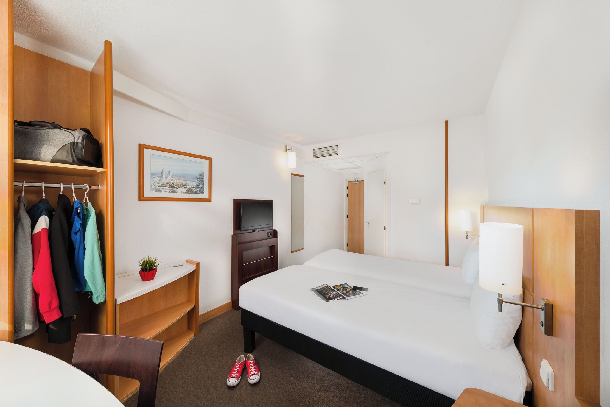Ein Hotelzimmer mit Bett, Schreibtisch, Stuhl, Fernseher, Kleiderschrank und einer kleinen Pflanze.
