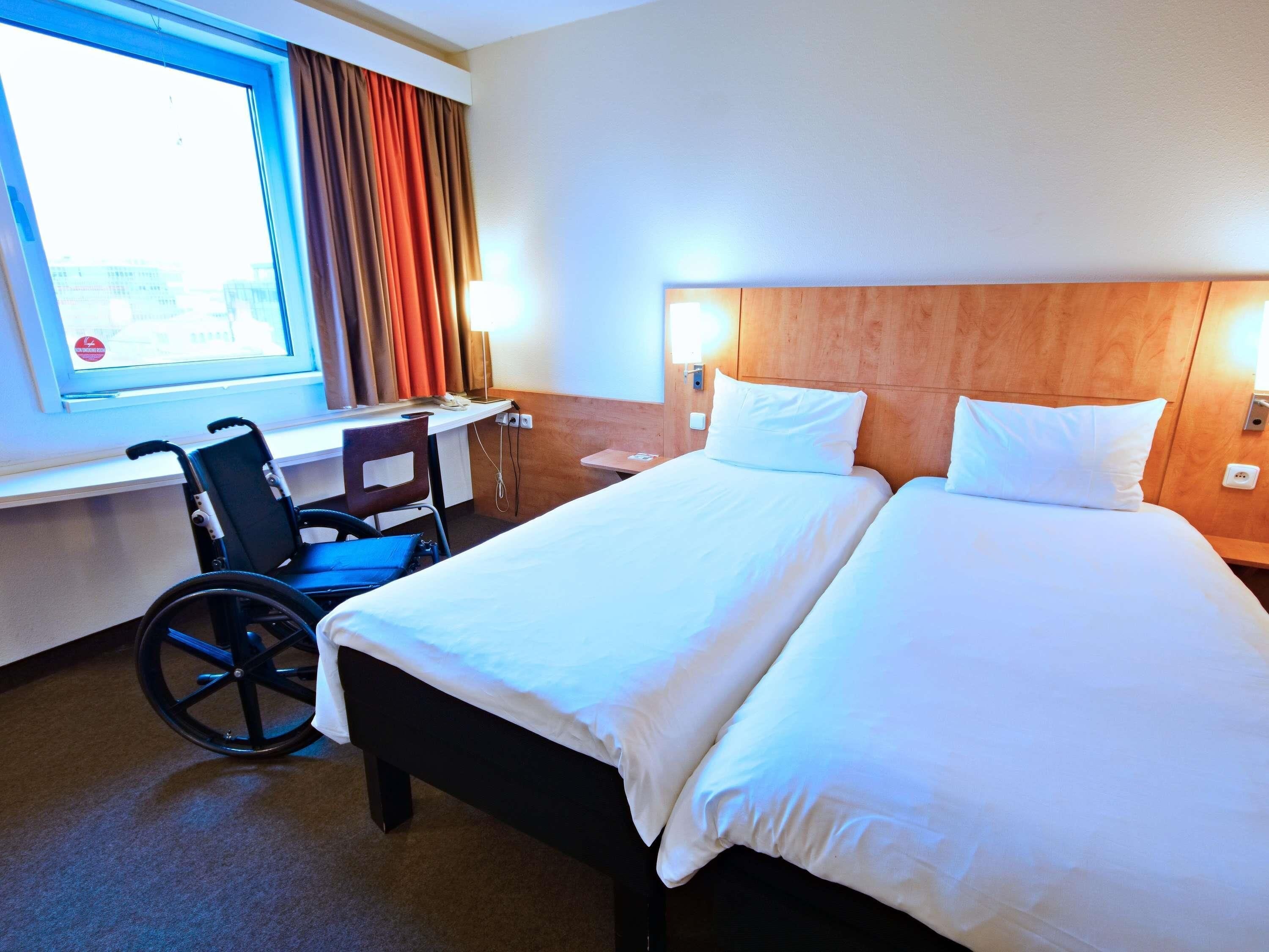 A hotel room with a wheelchair, a desk, and a bed with white linens.