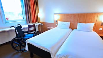 A hotel room with a wheelchair, a desk, and a bed with white linens.
