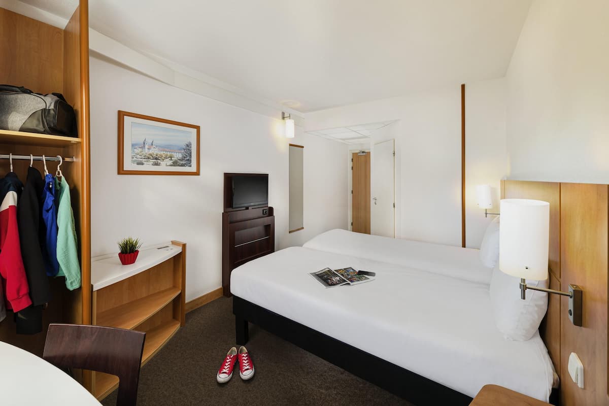 Une chambre d’hôtel comprenant un lit, un bureau avec une chaise, une télévision, un porte-manteau avec des vêtements et une petite plante.