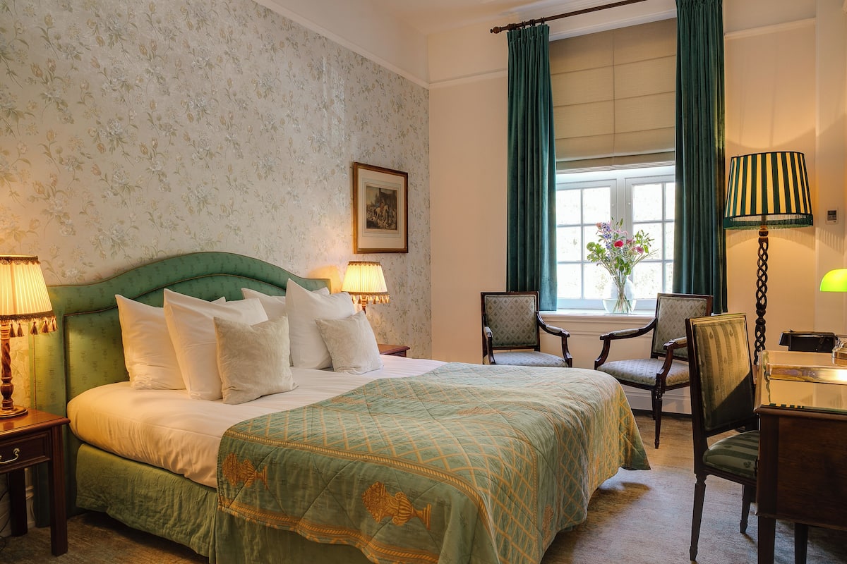 A hotel room with a large bed, two bedside lamps, a chair, a small table, and a window with curtains.